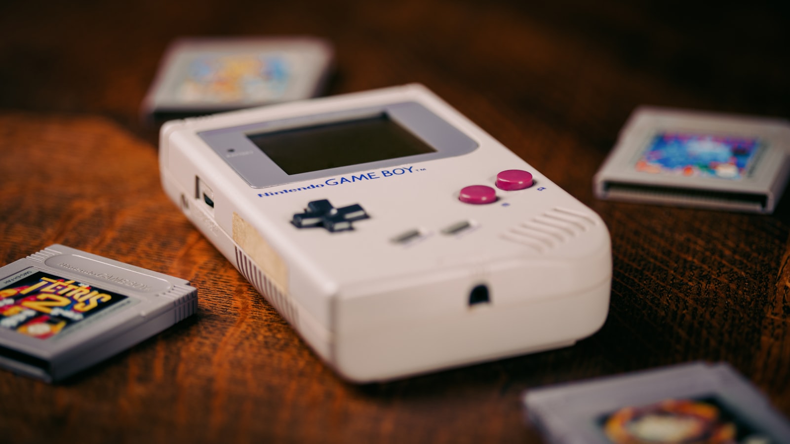 An original grey Nintendo Game Boy lying on a dark wooden table, surrounded by loose cartridges including a worn Tetris cassette.