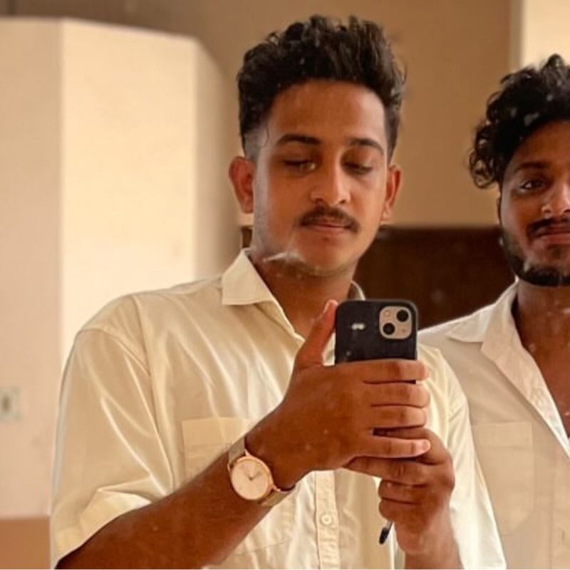 A mirror selfie of two young men in cream shirts in a plain unfinished room, one of them holding a phone up to take the picture.