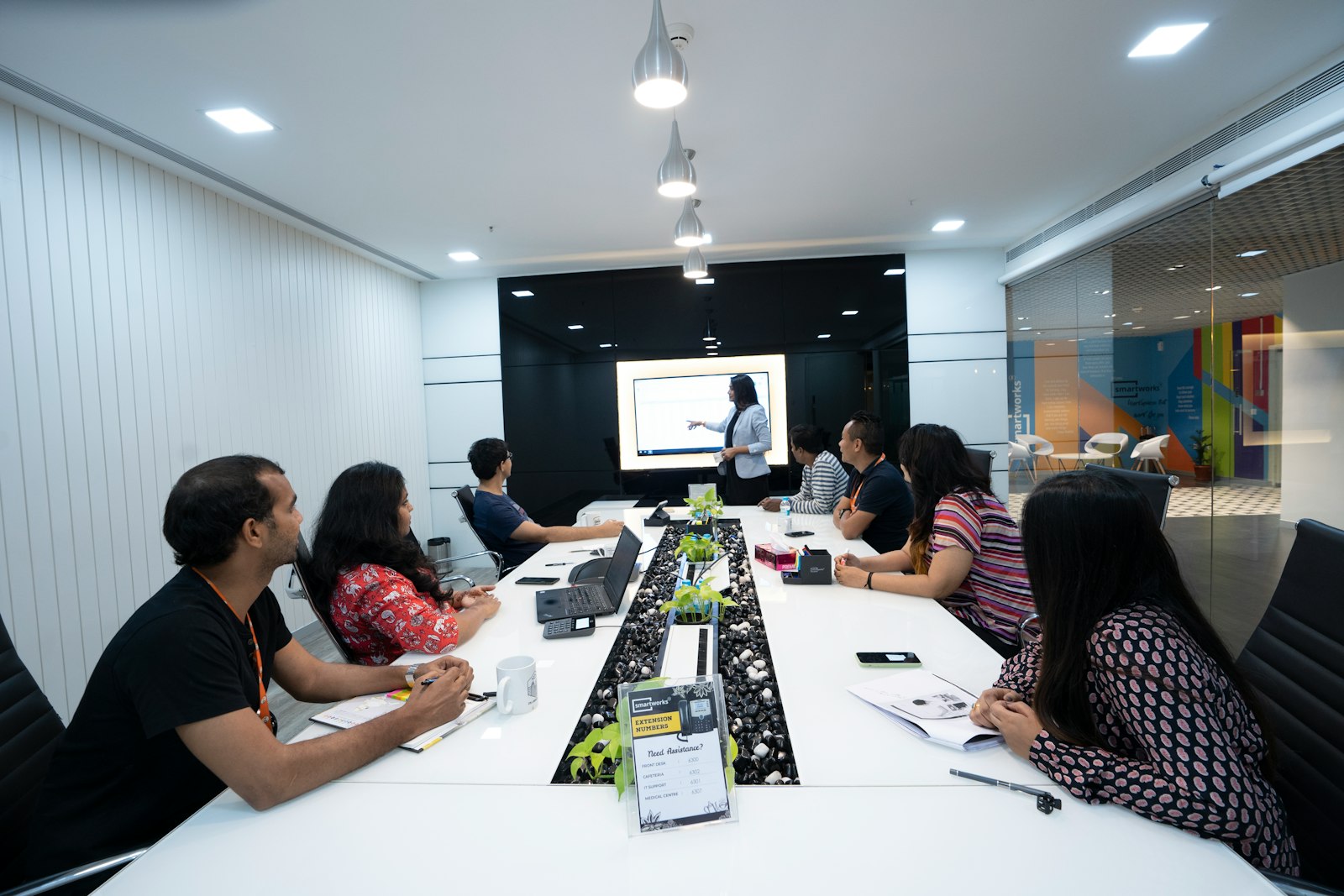 A team seated around a long white table in an office meeting room, laptops and notebooks open, listening to a colleague presenting at a screen.