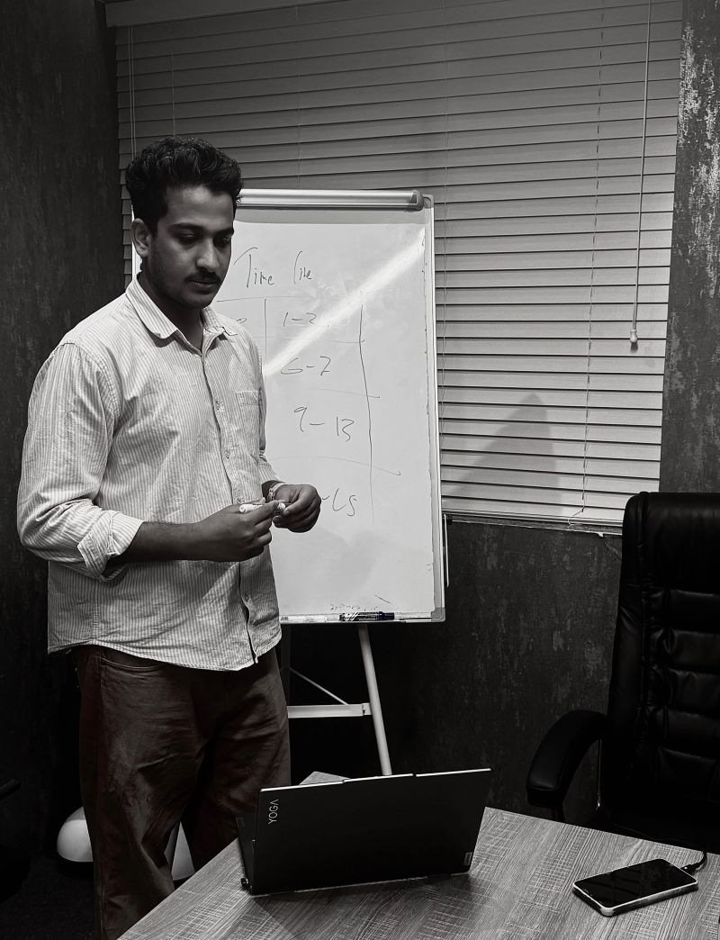 A black-and-white photo of a man standing beside a flip chart in a meeting room, capping a marker after writing a hand-drawn timeline with rows labelled 1-3, 6-7 and 9-13, an open laptop and a phone on the table in front of him.