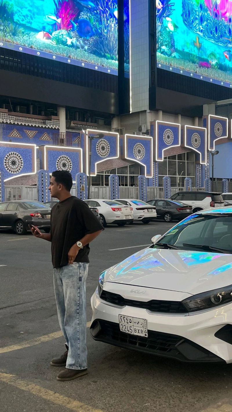 A man standing in a Saudi car park beside a white Kia with his phone in his hand, under a huge LED screen showing a coral reef above a shopfront tiled in blue and white.