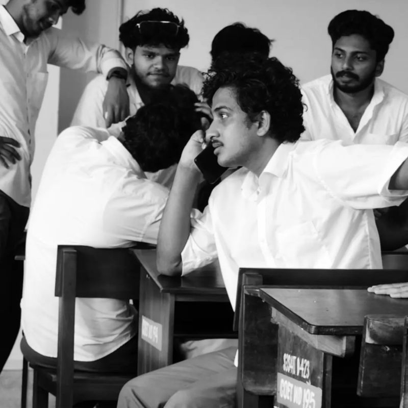 Black-and-white photo of Muhammed Nasvih on a phone call while seated among classmates at classroom desks.